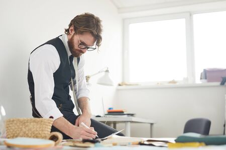 Serious designer sitting at his workplace and making notes in documents while working in the...