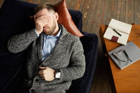 High angle view of young businessman lying on the sofa and closed his eyes with hand he tired of workの写真素材
