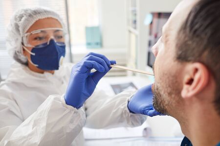 Mature man opening the mouth while doctor in protective clothing examining his throat during medical exam at hospitalの写真素材
