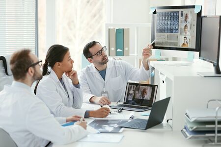Doctor pointing at computer monitor and discussing x-ray images with his colleagues during online conferenceの写真素材