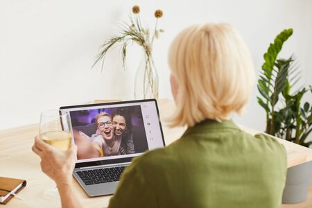 Rear view of woman with blond short hair holding glass of wine and celebrating the holiday with her friends online on laptopの写真素材