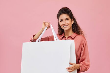 Portrait of young woman with curly hair holding big shopping bag and smiling at camera against the...