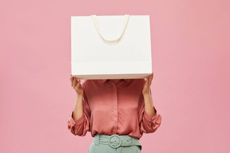 Young woman hiding before the big white shopping bag while standing against the pink backgroundの写真素材