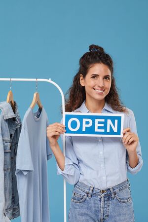 Portrait of young woman with curly hair smiling at camera and holding Open placard near the clothes on the rack isolated on blue backgroundの写真素材