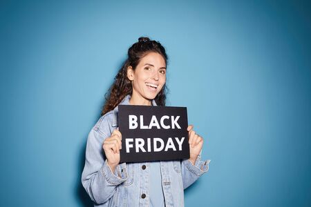 Portrait of young beautiful woman holding Black Friday placard and smiling at camera against the blue backgroundの写真素材