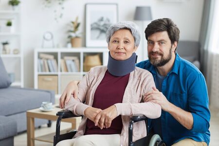 Portrait of senior woman in bandage on her neck sitting in wheelchair and looking at camera together with male nurse at homeの写真素材