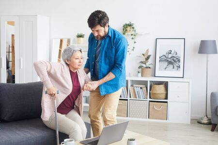 Man helping senior woman to stand up he support her while they are in the living room at home