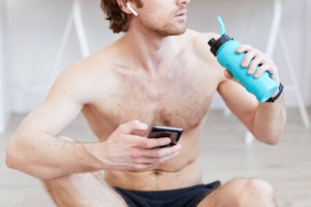 Shirtless muscular man drinking water and using his mobile phone after sports trainingの写真素材