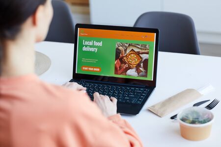 Rear view of young woman sitting at the table in front of laptop and ordering pizza onlineの写真素材