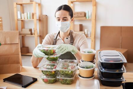 Young woman wearing protective mask packing healthy food in boxes she sitting at the table and working at officeの写真素材