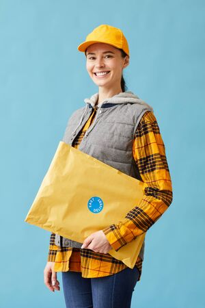 Portrait of young postwoman in uniform holding big parcel and smiling at camera isolated on blue...