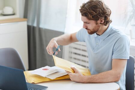 Young man sitting at the table in front of laptop and opening the paper envelopes at home