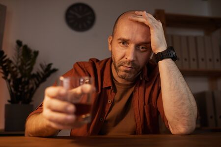 Portrait of sad mature man sitting at the table and looking at camera with alcohol drink