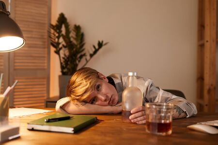 Portrait of young drunk woman lying on the table with bottle of whiskey near her she drinking...