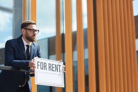 Serious mature realtor in suit holding placard and looking away while standing near the office buildingの写真素材