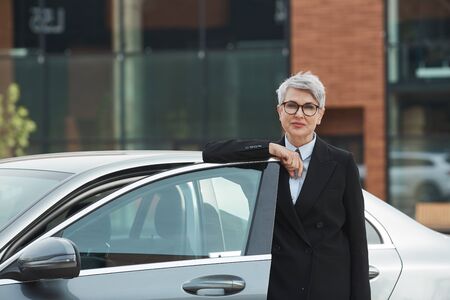 Portrait of mature businesswoman in eyeglasses standing near the car and looking at camera outdoorsの写真素材