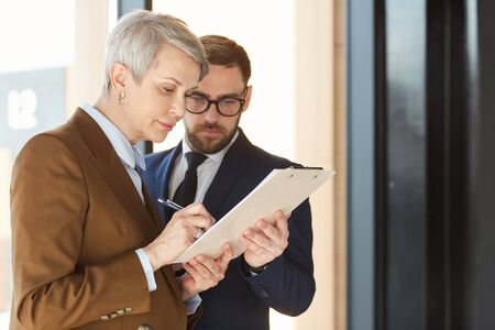 Serious mature businesswoman signing the business contract with businessman in suit standing near by...