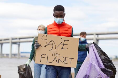 African young man in protective mask holding placard with other volunteers working in the background outdoorsの写真素材