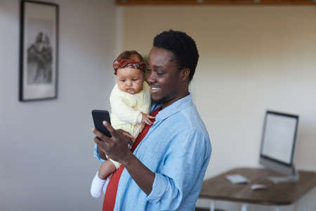 African smiling father holding baby girl on his hands and using his mobile phone at homeの写真素材