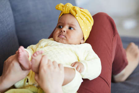 Beautiful baby girl lying on her mothers legs while they resting on sofa at home
