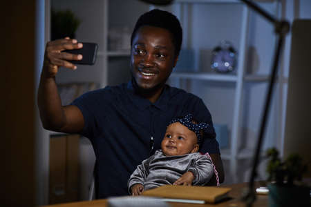 African happy father making selfie with his baby daughter on mobile phone while they sitting at the...