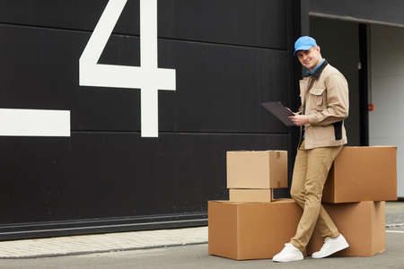 Portrait of young man filling document and smiling at camera while working in warehouse