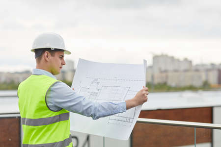 Serious engineer in work helmet examining the blueprint of new building while standing outdoorsの写真素材