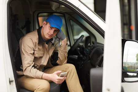 Portrait of delivery person in uniform holding mobile phone and looking at camera while sitting in the vanの写真素材