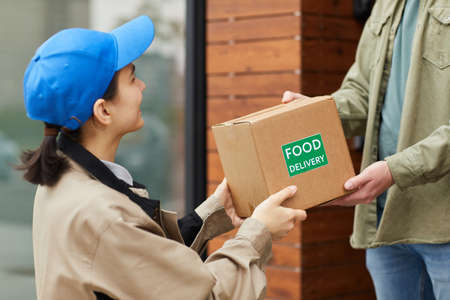 Young courier giving the cardboard box to man while they standing outdoors she delivering food