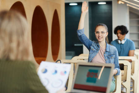 Content confident smart student girl in glasses raising hand to ask teacher at class in business...