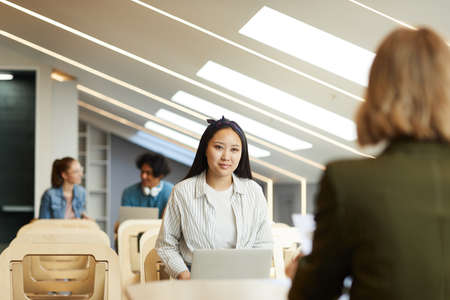 Content Asian student girl in shirt sitting in front of teacher and using laptop while passing...