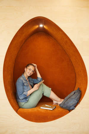 Portrait of smiling beautiful student girl sitting in egg-shaped armchair with satchel and notepad...