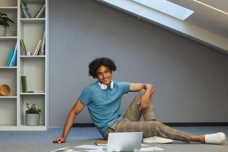 Portrait of smiling stylish student guy with disheveled hair sitting among papers and laptop in own...