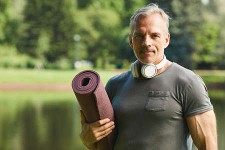 Portrait of handsome mature man with wireless headphones around neck holding rolled fitness mat in...