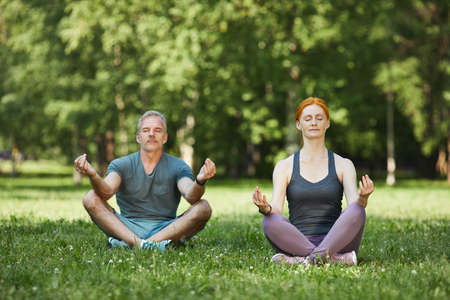 Peaceful mature couple concentrated on mind during meditation sitting with crossed legs in beautiful parkの写真素材