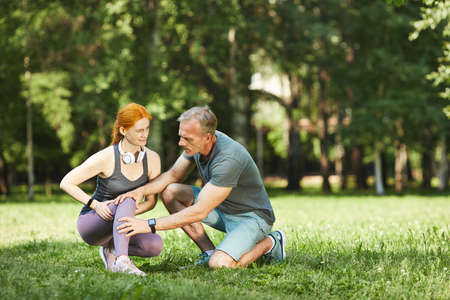 Careful mature fitness trainer checking knee of woman while she feeling pain after exercise in parkの写真素材