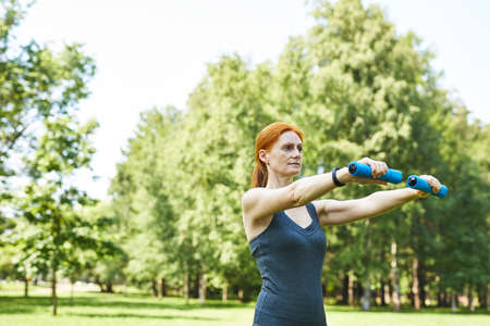Focused mature redhead woman using dumbbells for arm exercises in summer park