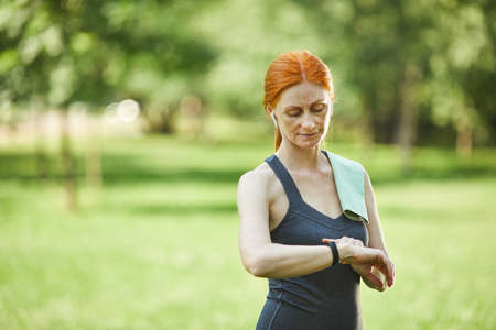 Attractive redhead mature woman using fitness tracker while checking pulse after outdoor training in parkの写真素材