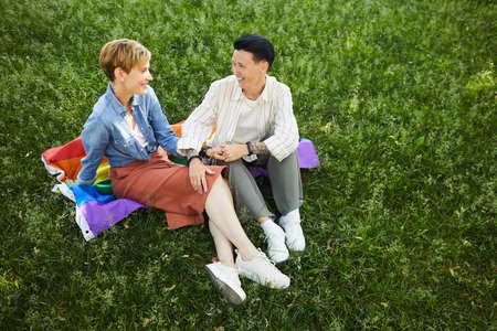 Happy lesbians sitting on green grass on colored flag and talking to each other during summer day