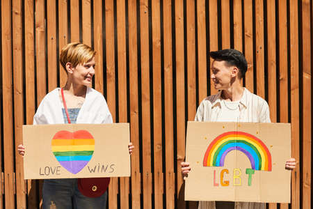 Young happy lesbians looking at each other and holding posters with LGBT signs while standing outdoorsの写真素材