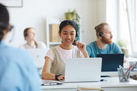 Asian young operator wearing headset sitting at workplace and using laptop she working at office...