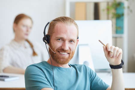 Portrait of young bearded man in headset smiling at camera while working at office