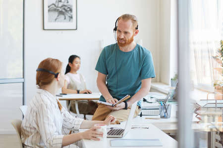 Bearded young customer service worker in headphones talking to his colleague while working in call center at officeの写真素材