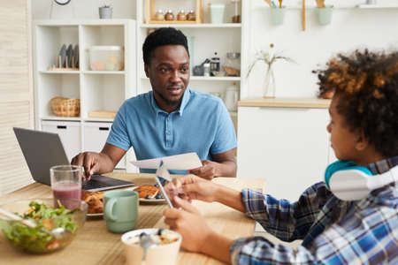 African young father sitting at the table in front of laptop and talking to his son while he playing...