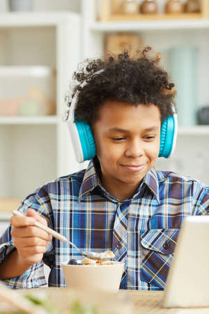 African little boy in headphones watching something on tablet pc while having breakfast at homeの写真素材