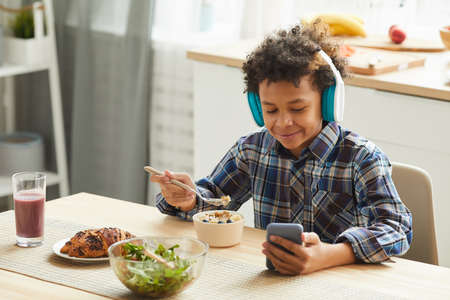 African schoolboy wearing headphones watching online lessons on his mobile phone and eating...