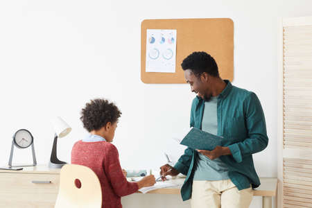 African boy sitting at desk and studying with his teacher at home teacher explaining the new...