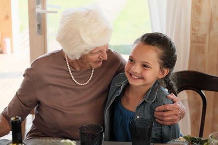 Happy grandmother embracing her smiling granddaughter while they sitting at the table and having...