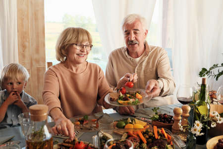 Happy senior woman putting vegetables on her husbands plate while they sitting at the table during...