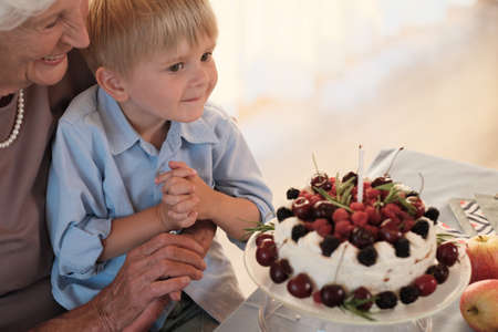 Little boy sitting on his grandmothers knees and blowing candle on his birthday cakeの写真素材